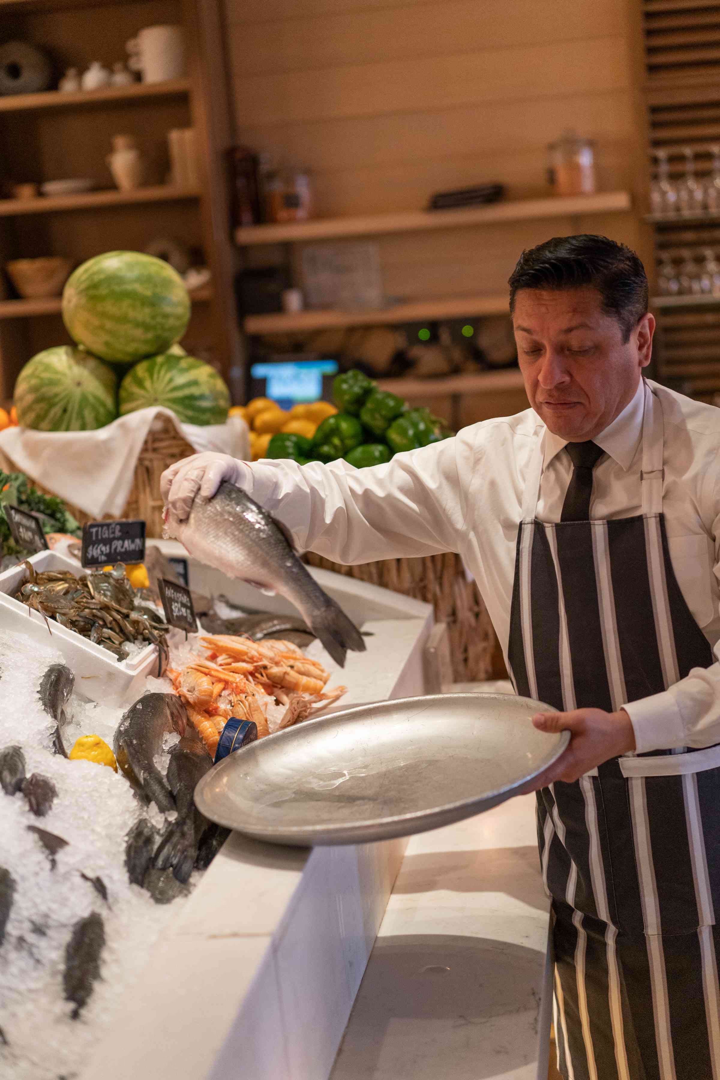 A chef prepares a large dish of seafood in a well-decorated kitchen with fresh ingredients around.