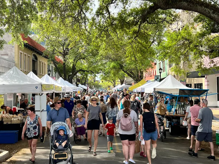 A bustling Bradenton, FL farmers market showcases a lively crowd exploring stalls under large trees, featuring fresh produce, artisanal goods, and local crafts on display.