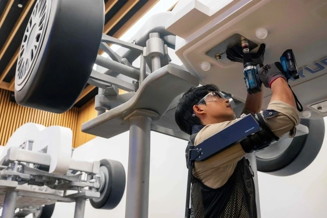 A person wearing a robotic exoskeleton uses a power tool to work on the underside of a large structure resembling a vehicle chassis, indoors. They are focused on their task, dressed in protective eyewear and gloves.