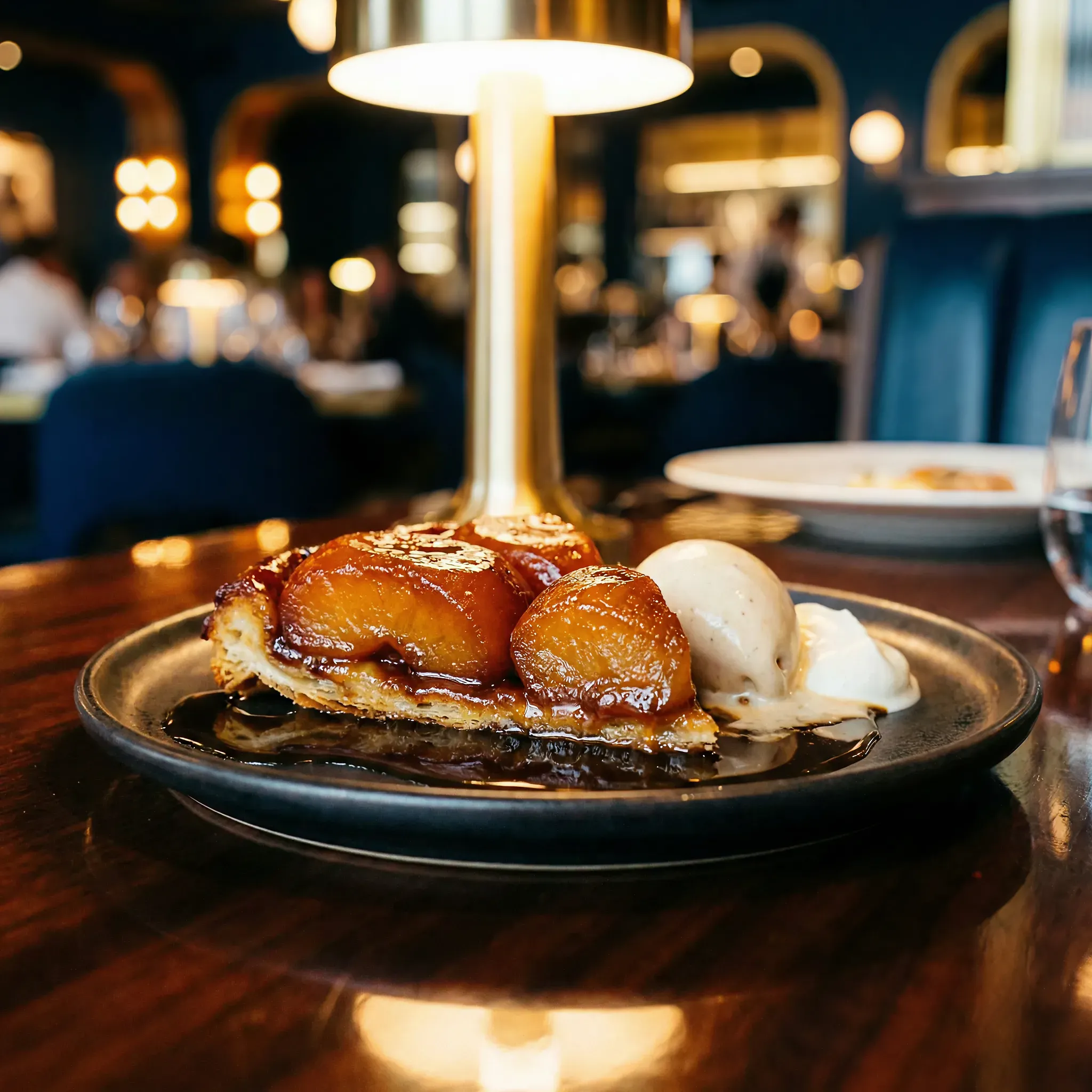 Caramelized apple tart with ice cream on a black plate, set on a wooden table in a warmly lit restaurant; cozy and inviting ambiance.