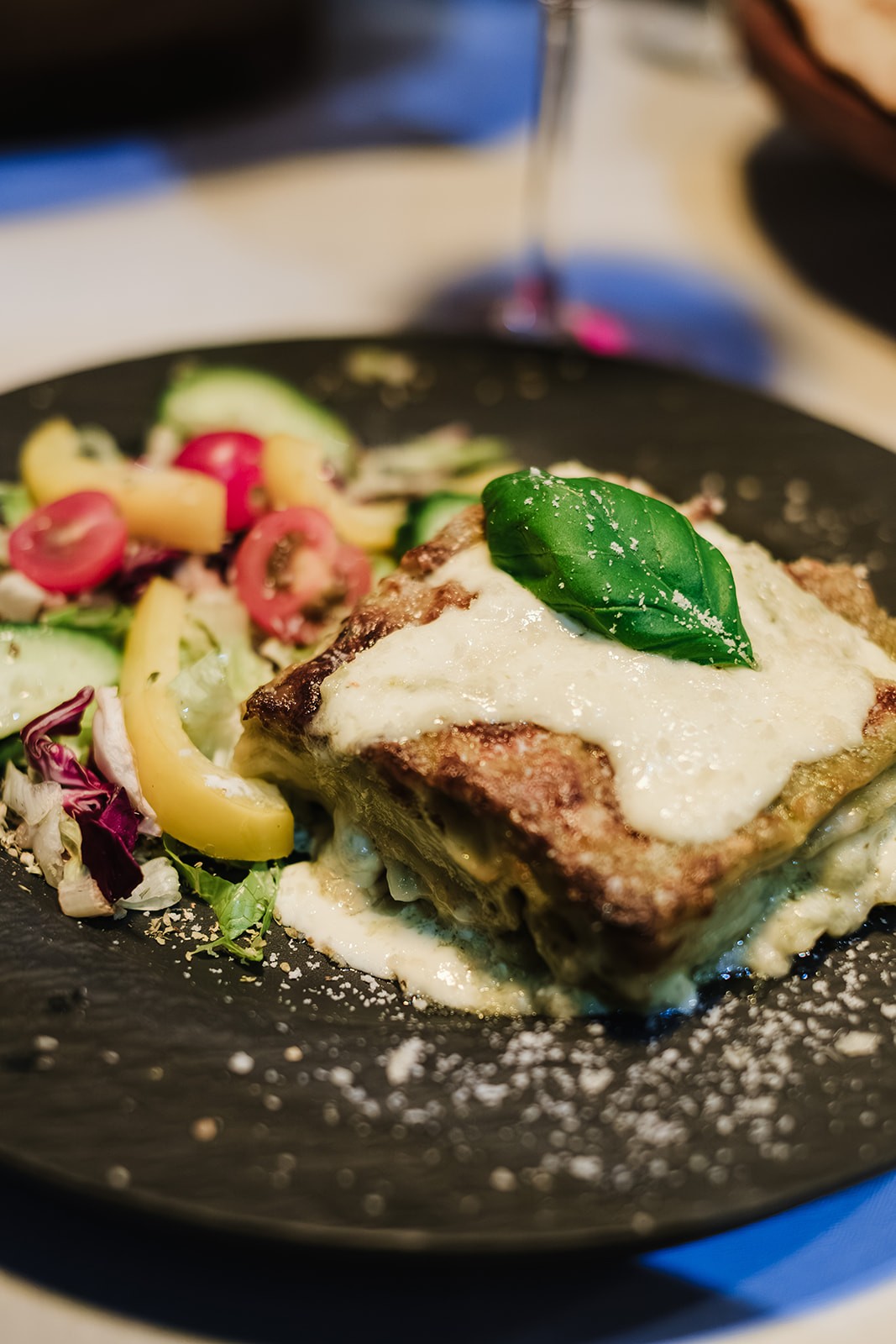 A close-up of a plated dish featuring a square portion of creamy lasagna topped with melted white sauce and a fresh basil leaf, served alongside a colourful side salad with sliced peppers, tomatoes and mixed greens on a dark plate.