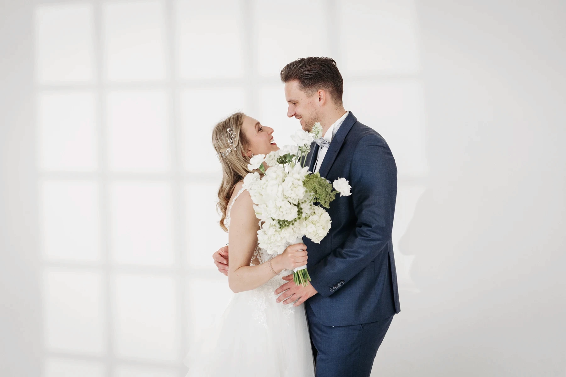 Braut in weißem Spitzen-Hochzeitskleid steht vor einem Fenster im Fotostudio Erfurt, mit sanftem Licht und Schatten.