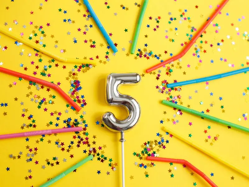A number 5 balloon with a metallic finish takes center stage on a bright yellow background, surrounded by a burst of multicolored star confetti and a fan of colorful party straws radiating outward. The scene exudes a festive and cheerful vibe, perfect for celebrating a 5-year milestone.