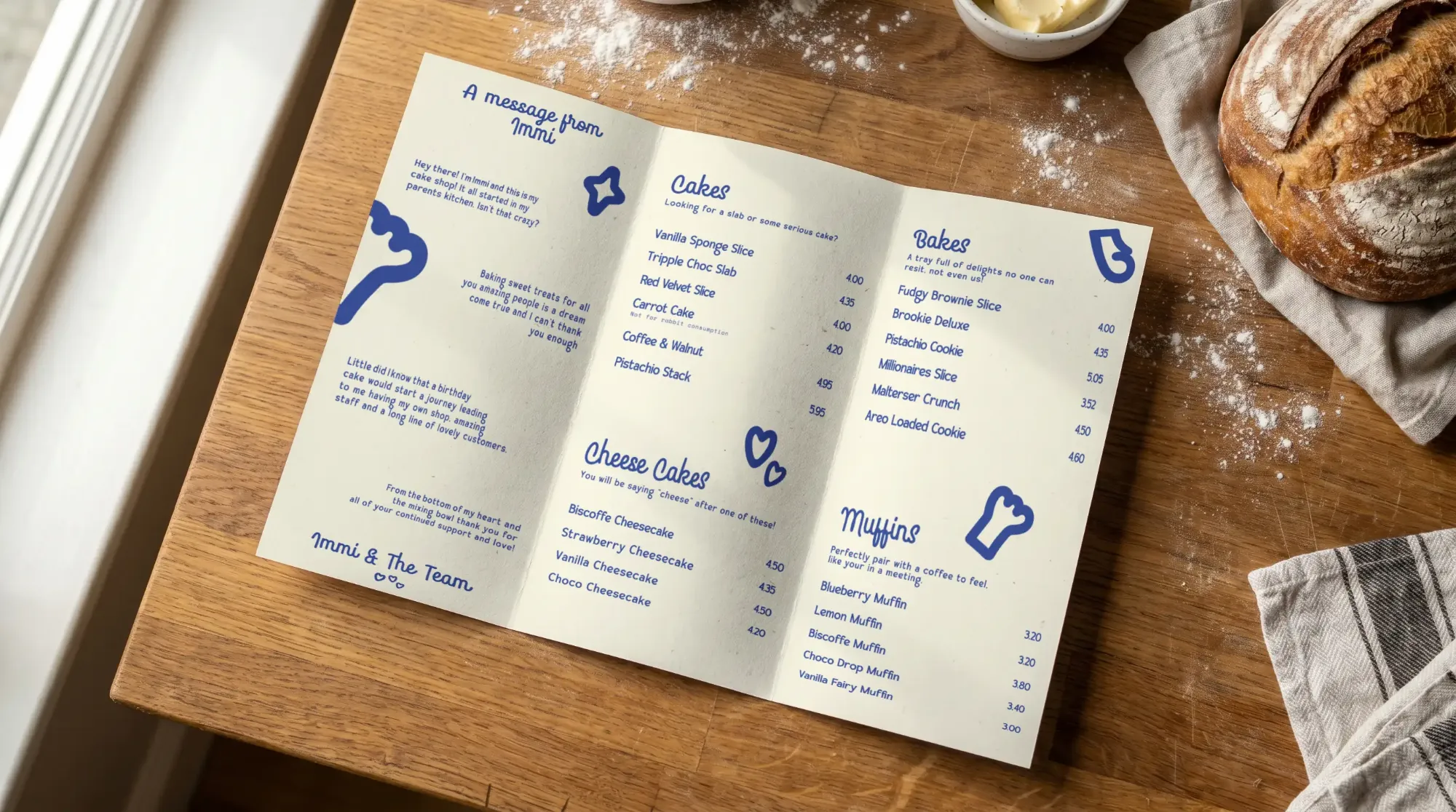Folded Immis menu listing cakes, bakes, cheesecakes and muffins on a floury wooden table