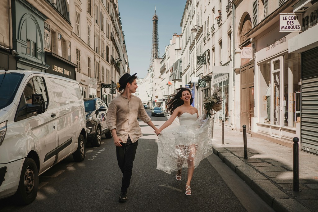 Laughing bride and groom running hand in hand down a sunny Paris street with the Eiffel Tower rising between the buildings