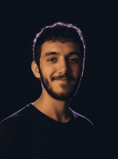 Retrato de Luigi Carvalho, um jovem adulto com barba curta e cabelo castanho, sorrindo levemente em um fundo escuro com iluminação suave ao redor da cabeça.