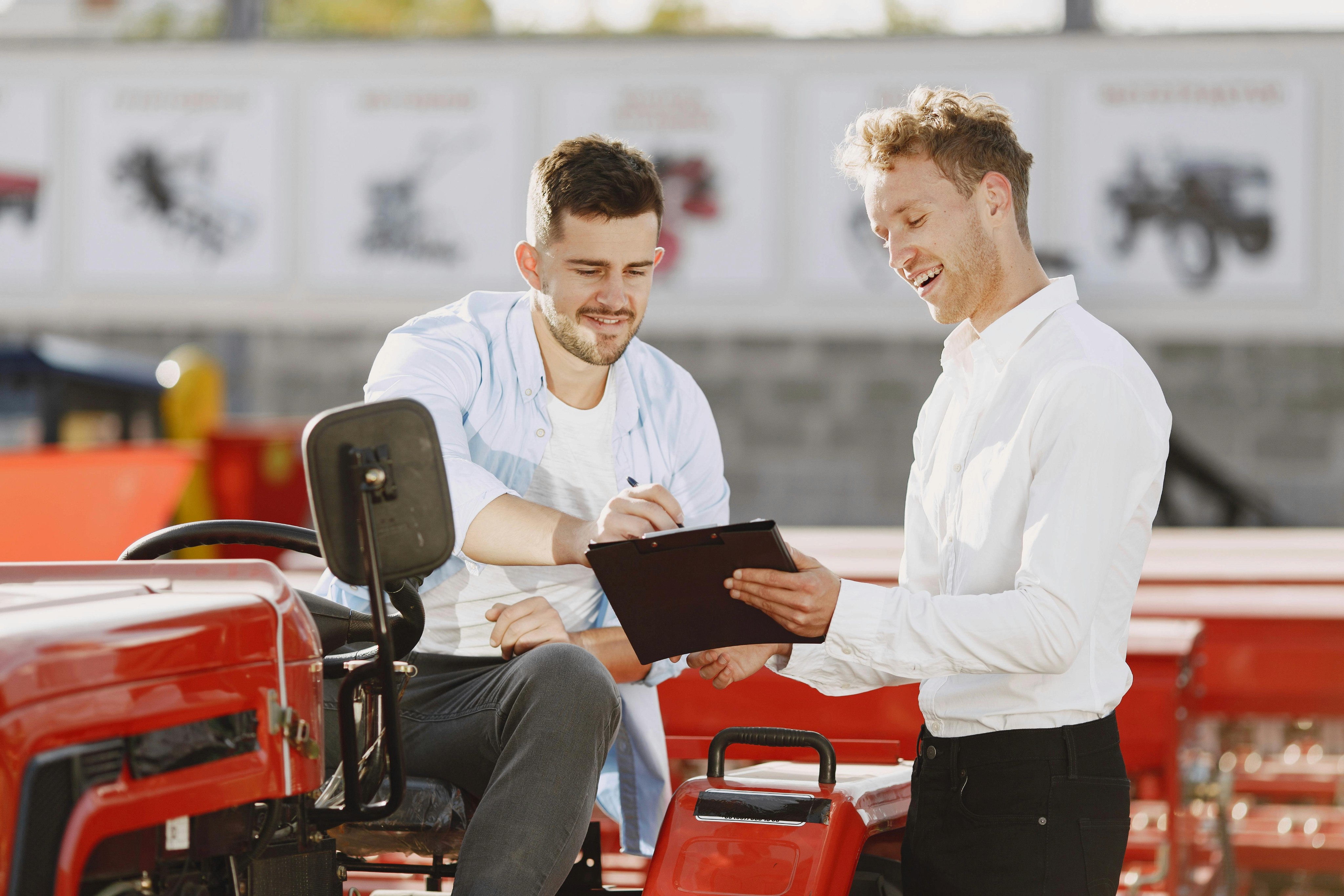 Two smiling men at an outdoor farm equipment dealership; one on a red tractor signs a clipboard held by the other.