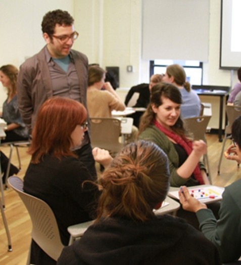 A person wearing glasses standing near a group of people that sit at a table and discuss.