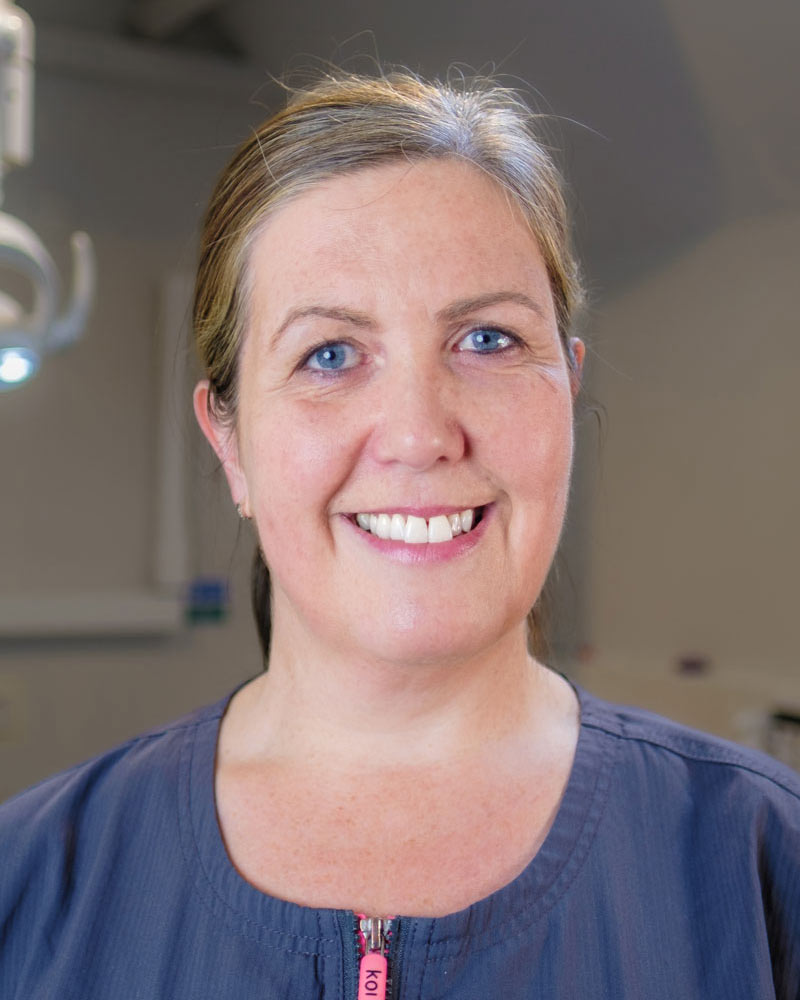 A portrait of Karen, a Dental Nurse at Cricklade Dental Practice, smiling and wearing a dark grey scrub top with a pink zipper pull and yellow embroidered text that reads "KAREN" on the left and "Cricklade Dental Practice" on the right. She is standing in a dental surgery with a dental light visible in the background.