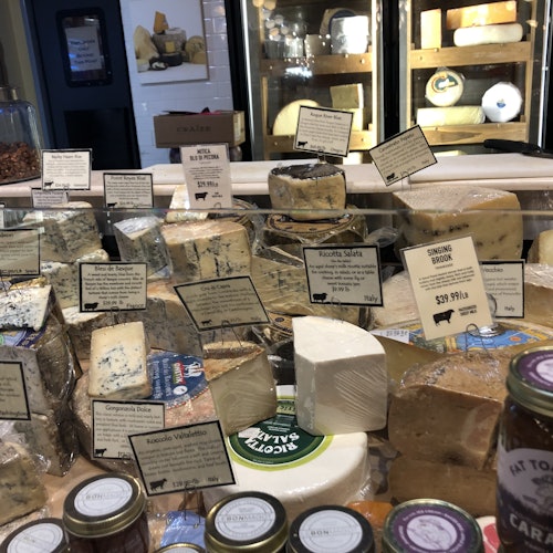 A display of various cheeses with labels, including blue cheese and gouda, in a deli setting with jars of condiments in the foreground.
