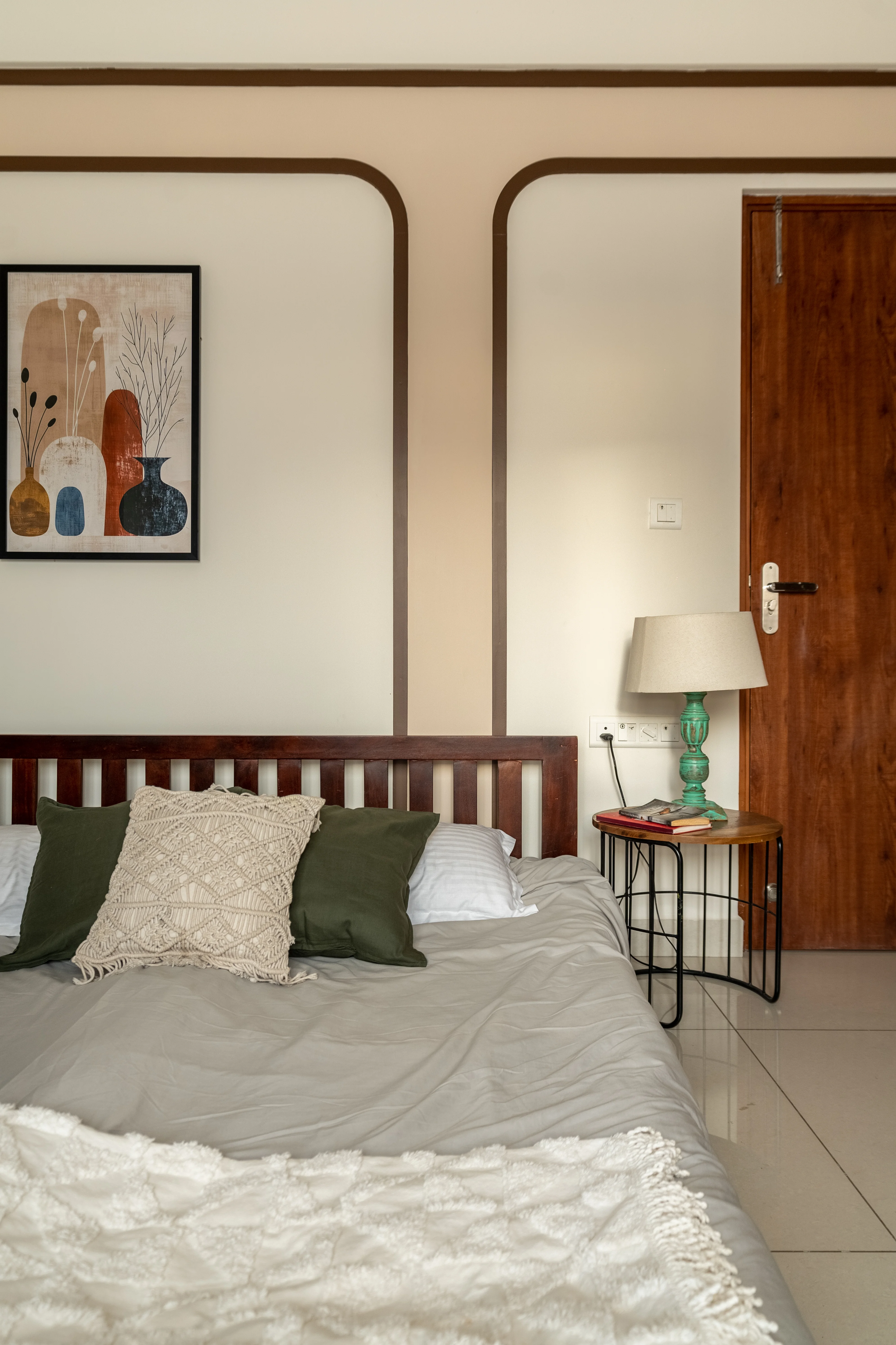 A bedroom with a wooden bed, featuring a gray headboard and a variety of pillows. A small bedside table holds a lamp and a book. The wall behind the bed is painted white with a decorative border.