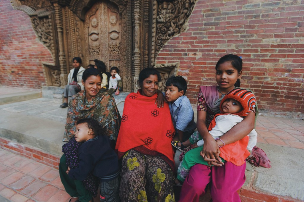 The image features a group of women and children sitting together outside a building with intricate wooden carvings on the door, likely in a South Asian setting. The women are dressed in traditional, colorful clothing, and they appear to be enjoying a relaxed moment together. The children, some sitting on laps and others standing nearby, add to the warm, communal atmosphere. The brick wall and carved doorway provide a rich, cultural backdrop, highlighting the traditional architecture and familial bonds.