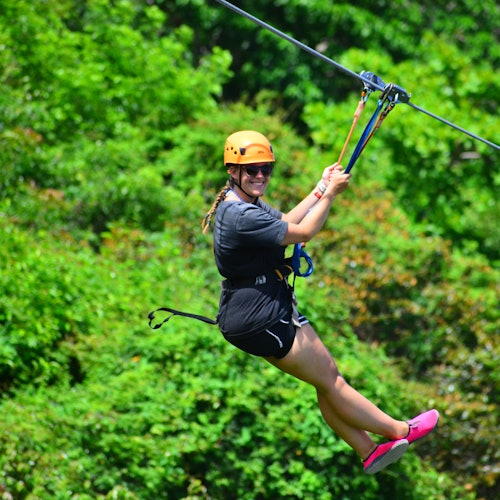 Persona sonriendo y haciendo tirolesa a través de un exuberante bosque verde, usando un casco, arnés y zapatos rosas.