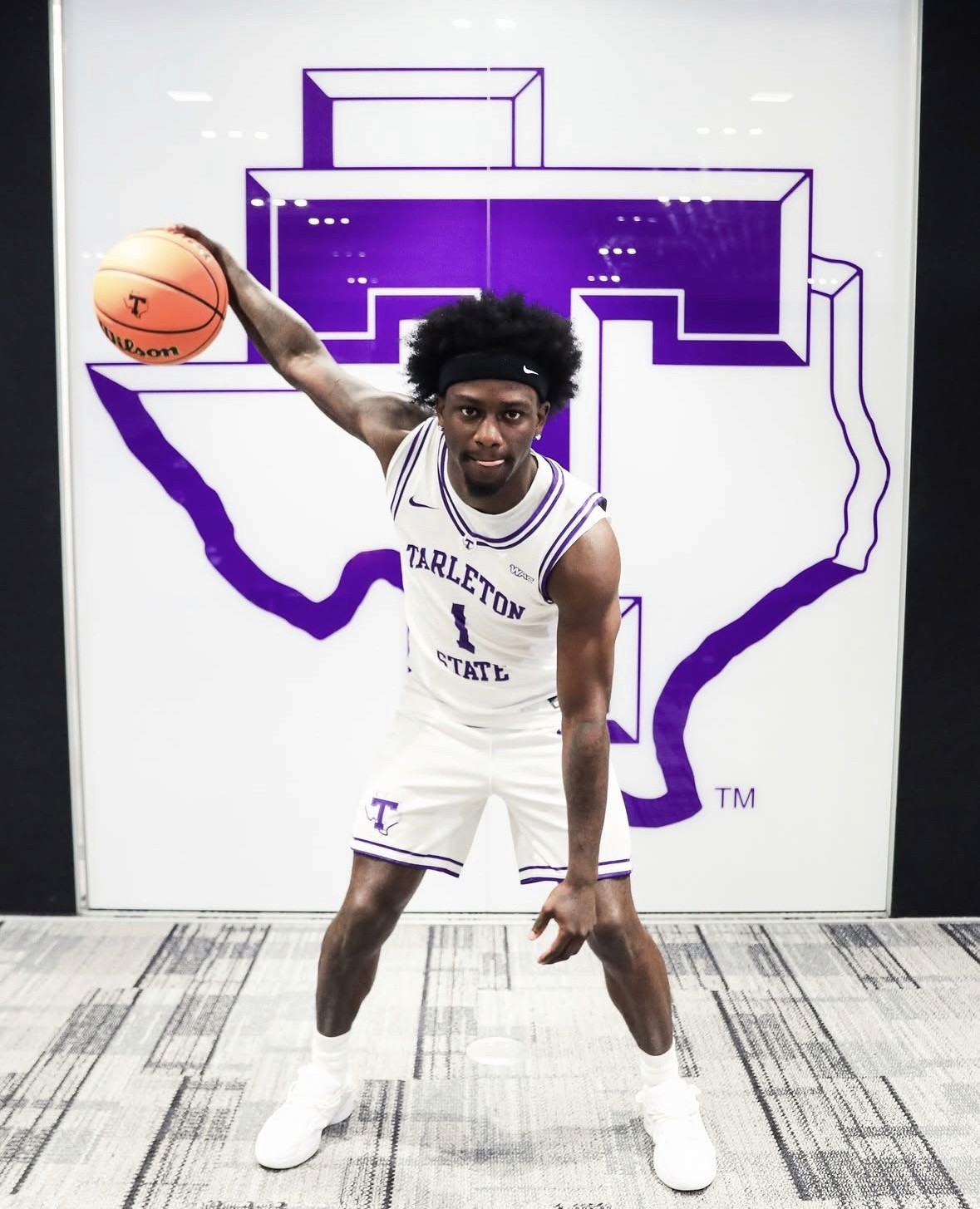 Caleb Blackwell in a Tarleton State uniform poses with a basketball, one arm extended. Behind him is a large purple outline of Texas with a Tarleton “T” logo. The player is on a carpeted floor, facing the camera.