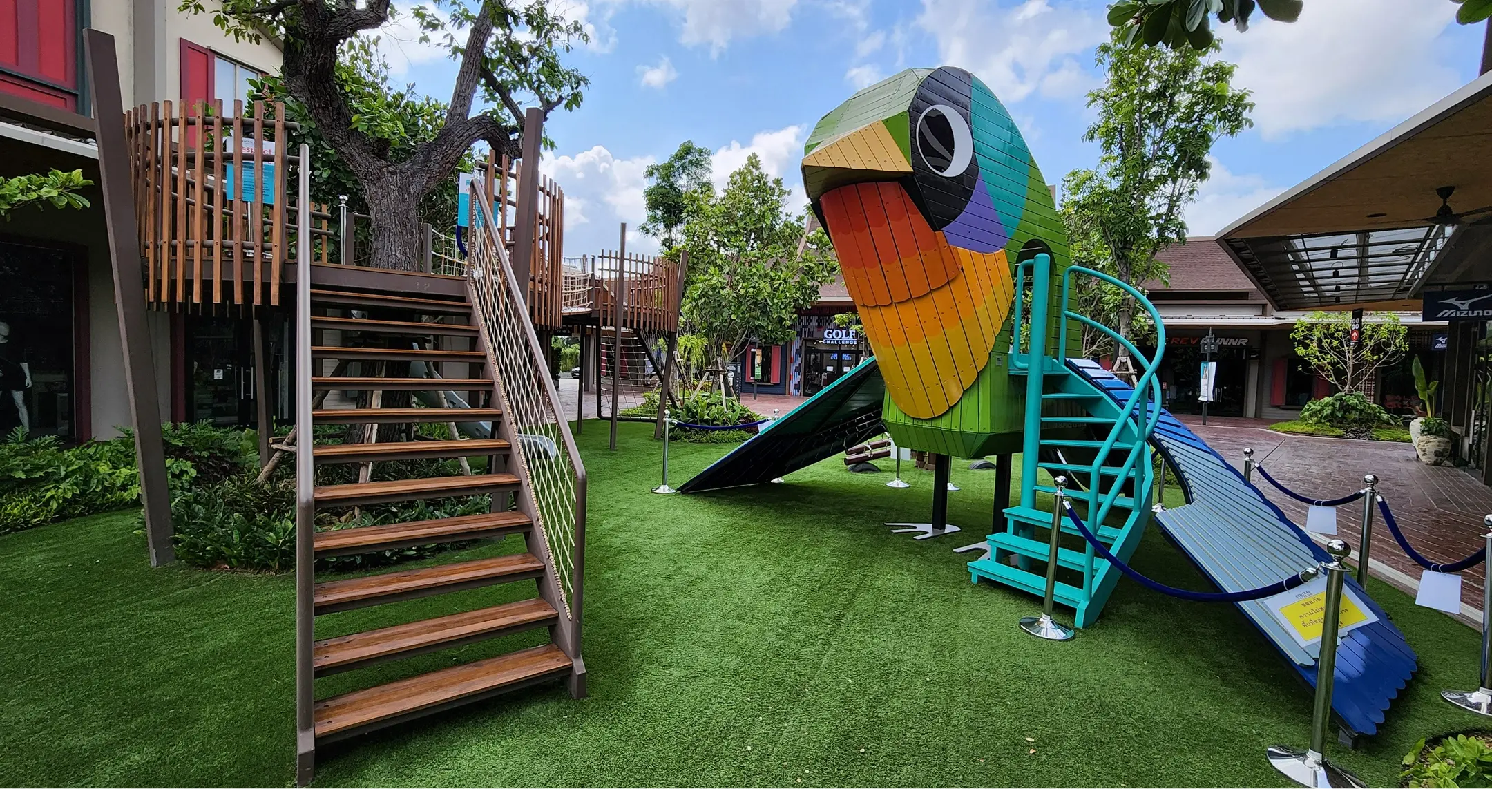 สนามเด็กเล่นที่มีสไลด์ของนกแก้วน่ารักและบ้านต้นไม้อันน่าหลงใหล มีสีสันสดใส บันไดไม้ พื้นหญ้าเขียวขจี ต้นไม้ และบรรยากาศที่มีชีวิตชีวา