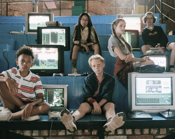 A group of five teenagers dressed in 90s style sits among a cluster of old CRT computer monitors displaying retro graphics.
