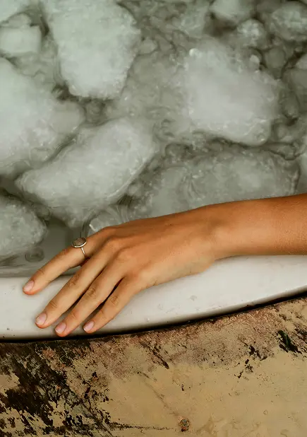 A hand with a ring rests on the edge of a bathtub filled with ice cubes, creating a contrast between the warm skin tone and the frosty ice.