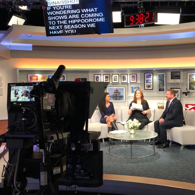 Three people sit and talk on a brightly lit TV studio set with cameras and a teleprompter visible in the foreground.