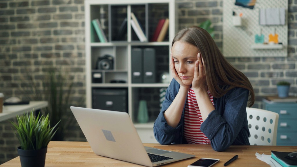 A young woman in a striped shirt and denim jacket sits at a wooden desk, looking tired and stressed as she stares at her laptop in a modern home office setting.