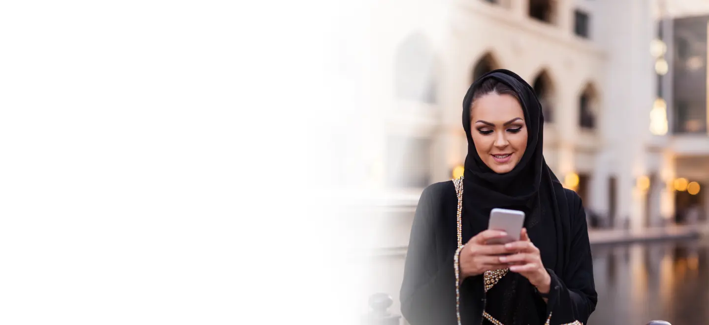 A woman in a black abaya smiling while using a health app on her smartphone outdoors, symbolizing the accessibility and convenience of digital health solutions for managing chronic conditions.