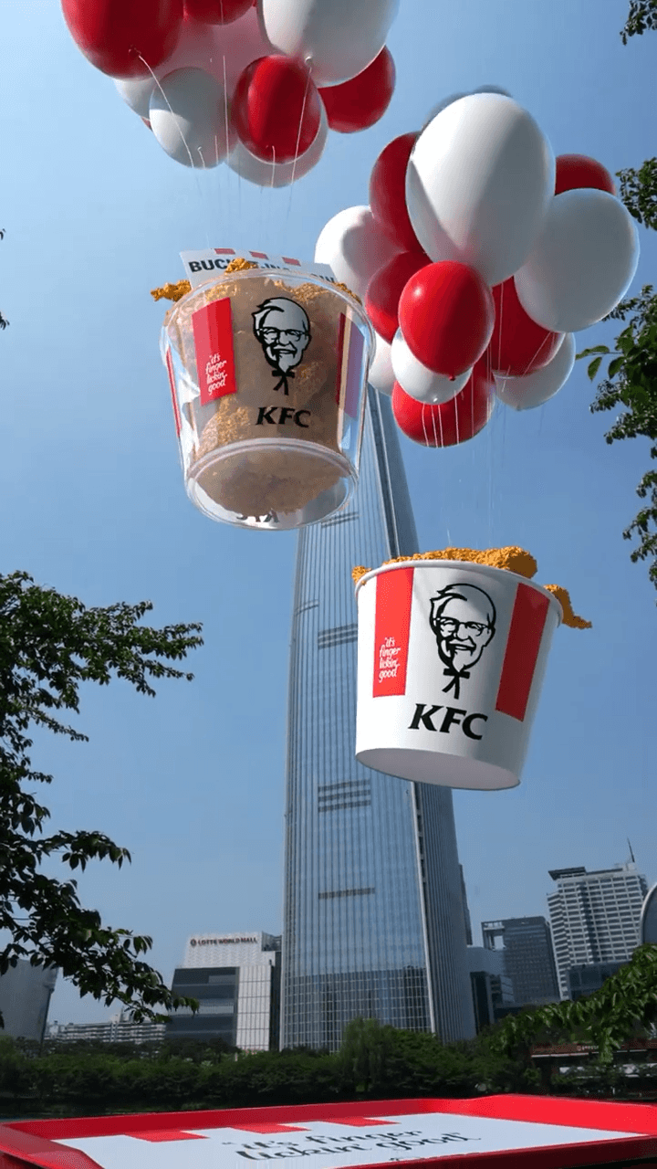롯데월드타워 앞에서 빨간색과 흰색 풍선에 매달린 KFC 치킨 버킷이 하강하는 FOOH 장면