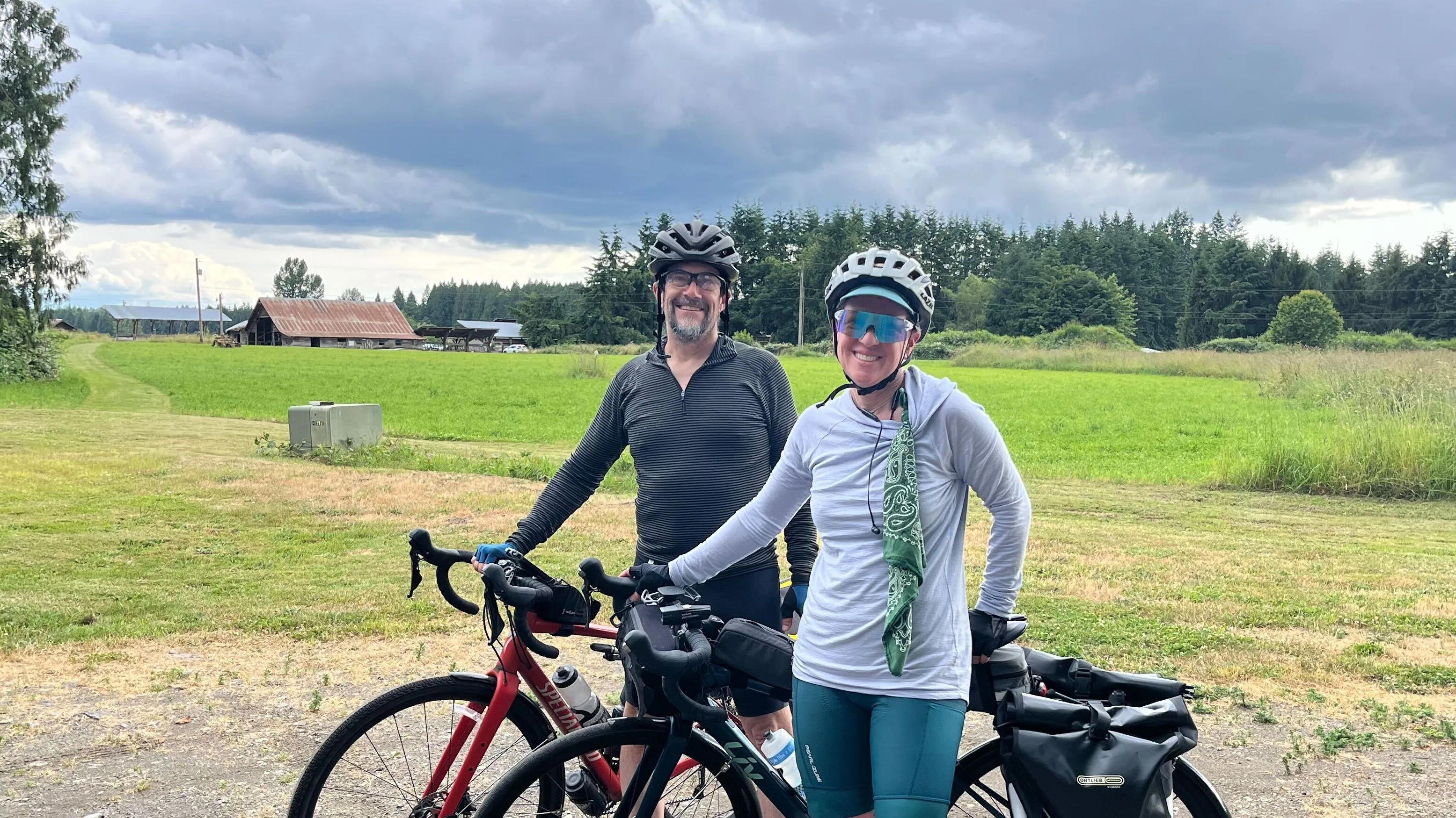 Two cyclists stand with touring bikes in an open field at Rooted Northwest, with barns, farmland, and forested hills in the background.