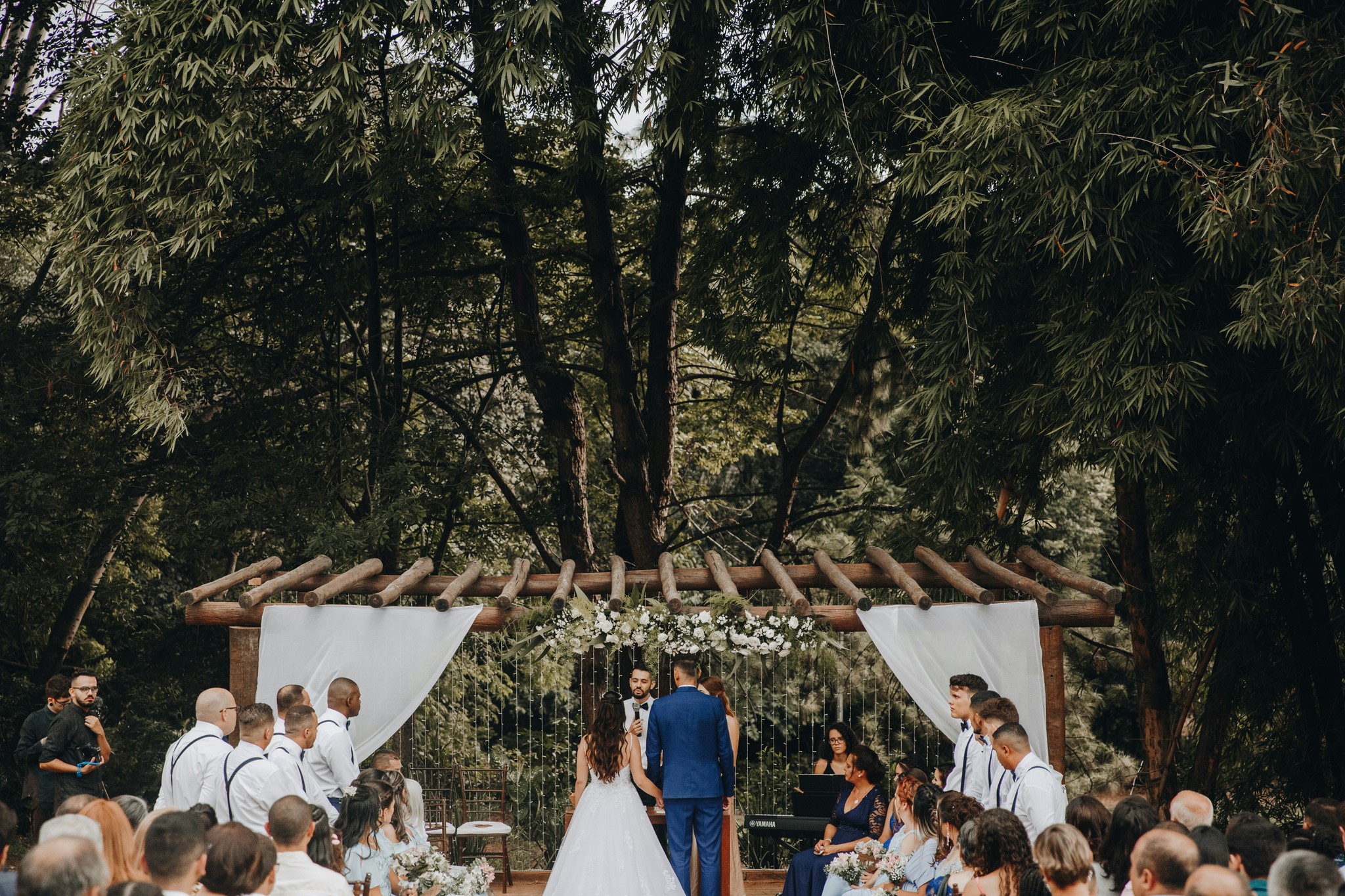 Bride and groom exchanging vows under a rustic wooden pergola with white draping surrounded by guests and lush forest trees
