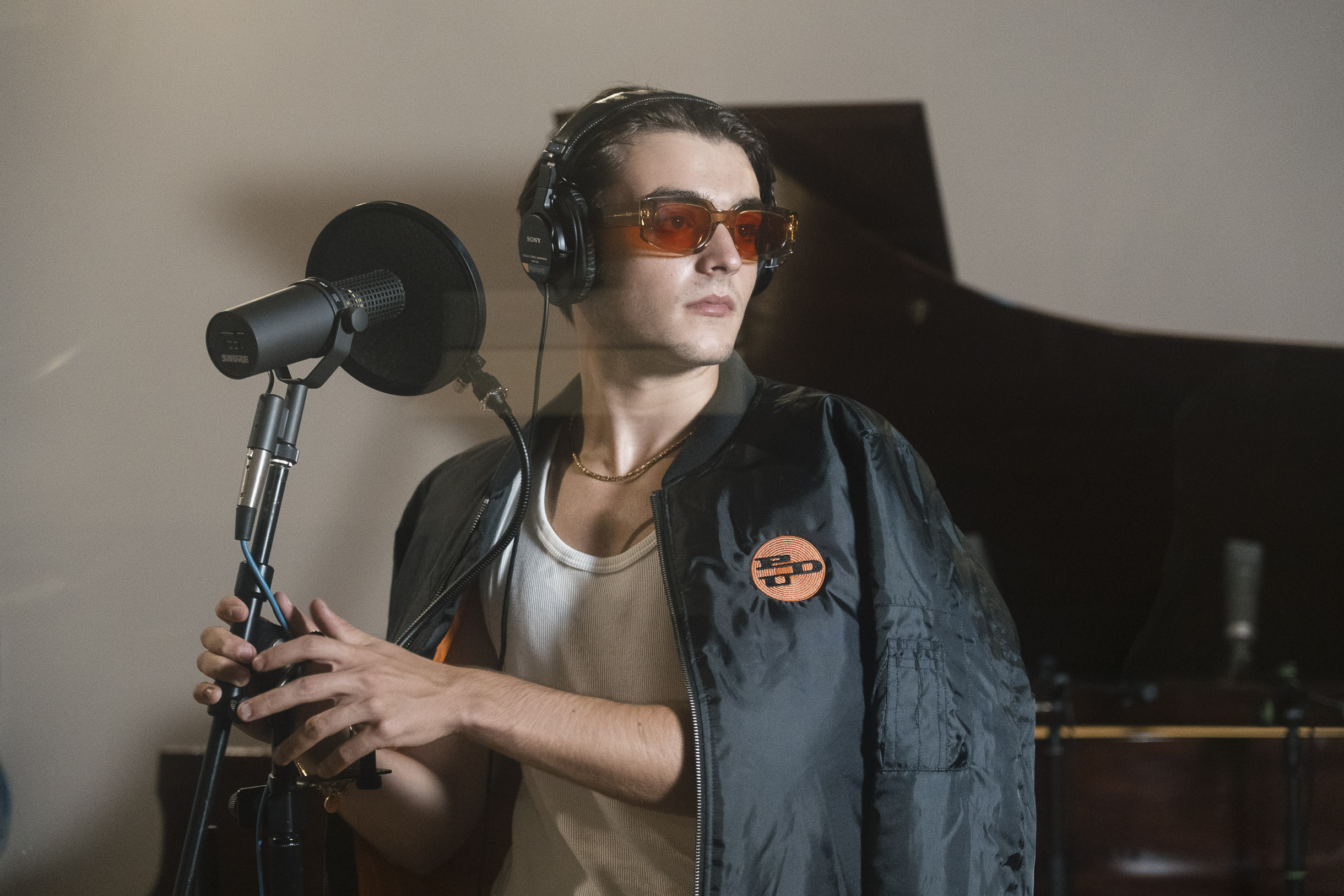 Photo dans un studio de musique de l'artiste italien Ghis tenant un micro sur pied et portant une veste bombers à l'effigie de PDU