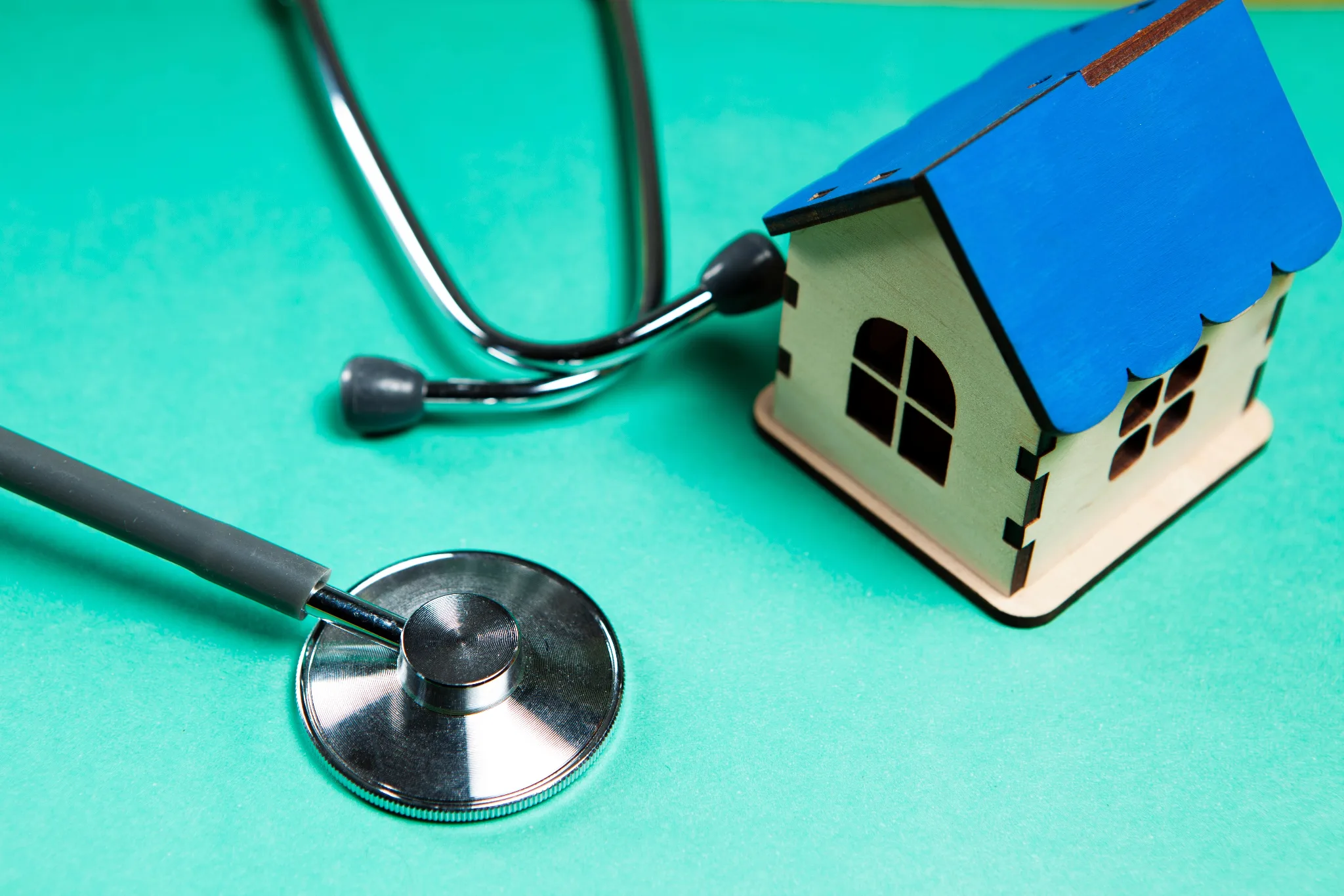 A stethoscope and miniature house model with a blue roof sit on a turquoise surface, symbolizing home health care or medical property inspection.