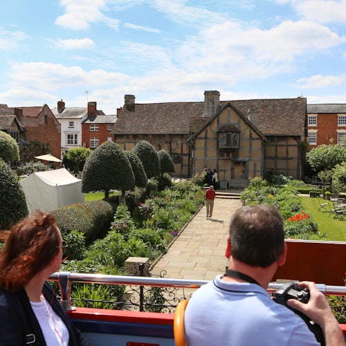 Zwei Touristen beobachten an einem sonnigen Tag von einem offenen Bus aus ein historisches Haus mit einem gepflegten Garten.