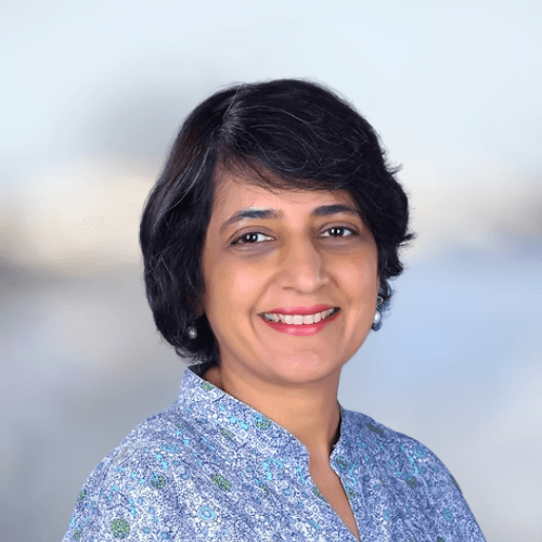 A smiling man with curly hair is sitting at a desk, looking at the camera, with a laptop and a lamp in the background.