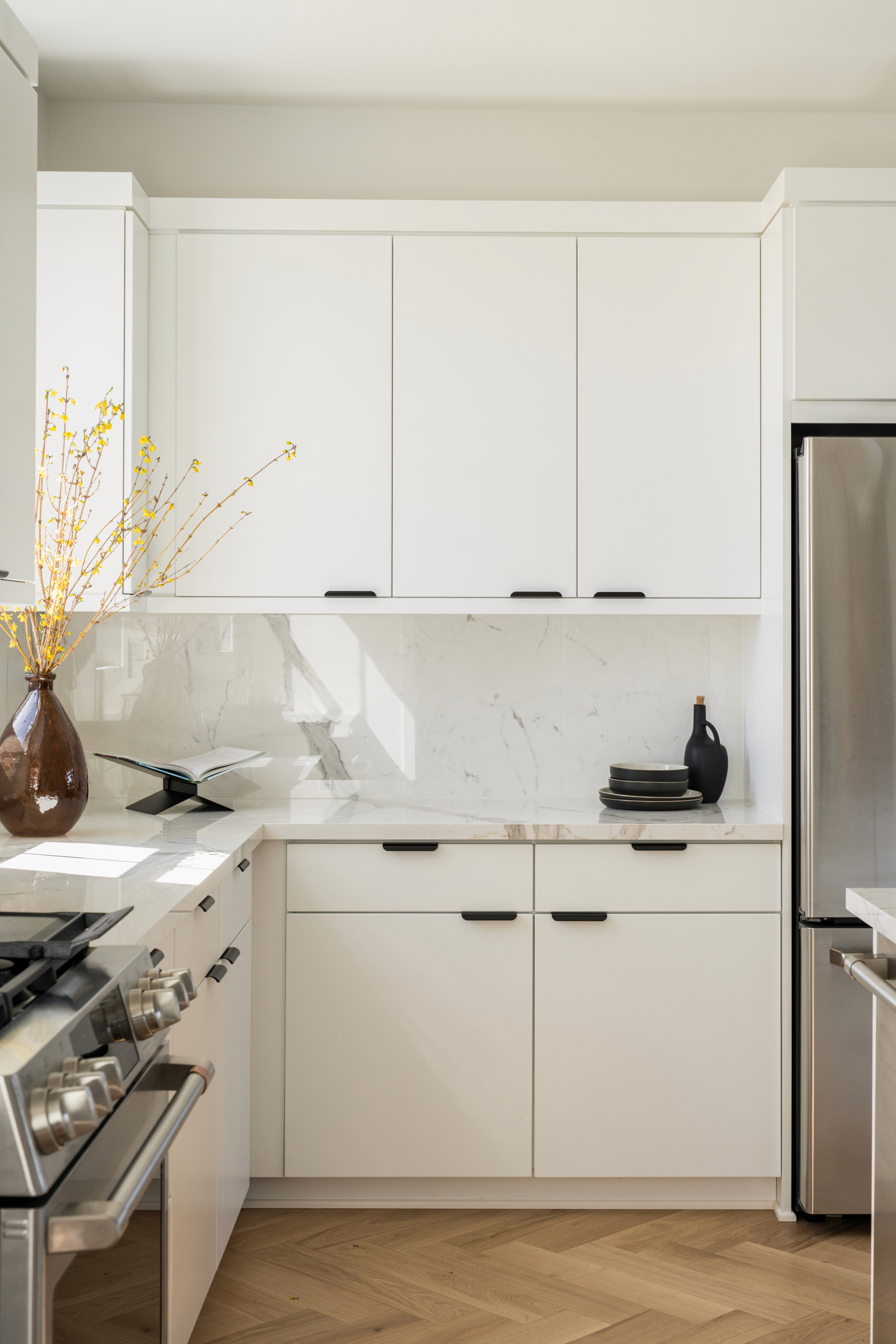 Marble kitchen countertop and backsplash with flat white cabinets, black hardware, and amber glass vase with dried natural flowers, stainless steel appliances and wood chevron flooring