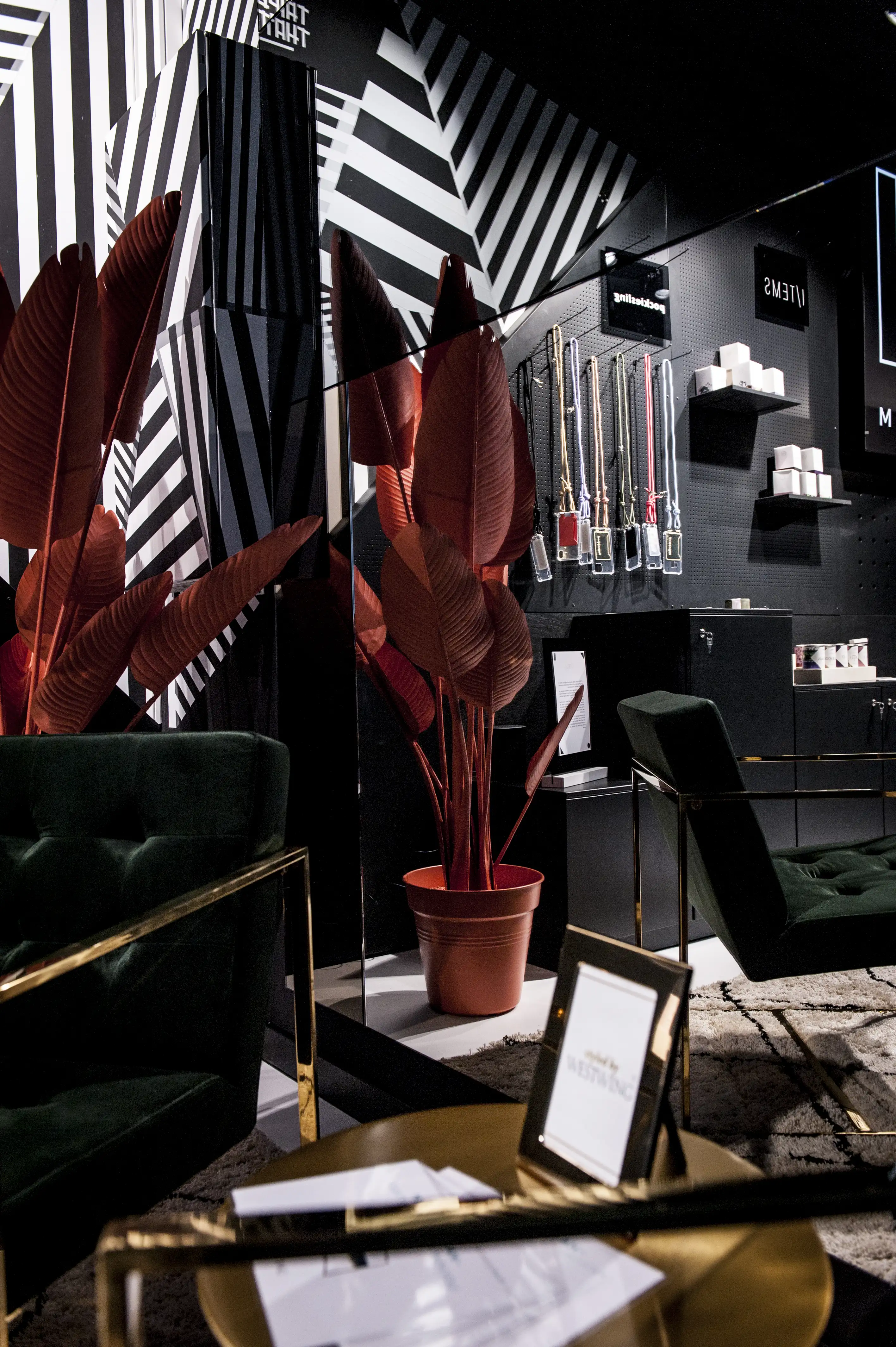 Dark brand area at FREIRAUM SHOP with black pegboard walls, hanging product tubes, white boxes on shelves, green velvet armchairs, gold side tables and a red banana plant.