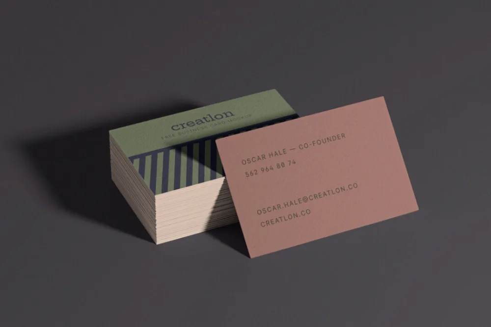 Business card mockup featuring a stack of cards with a green design and stripes, alongside a single card in earthy pink with minimalist typography. Set against a dark, softly lit background.