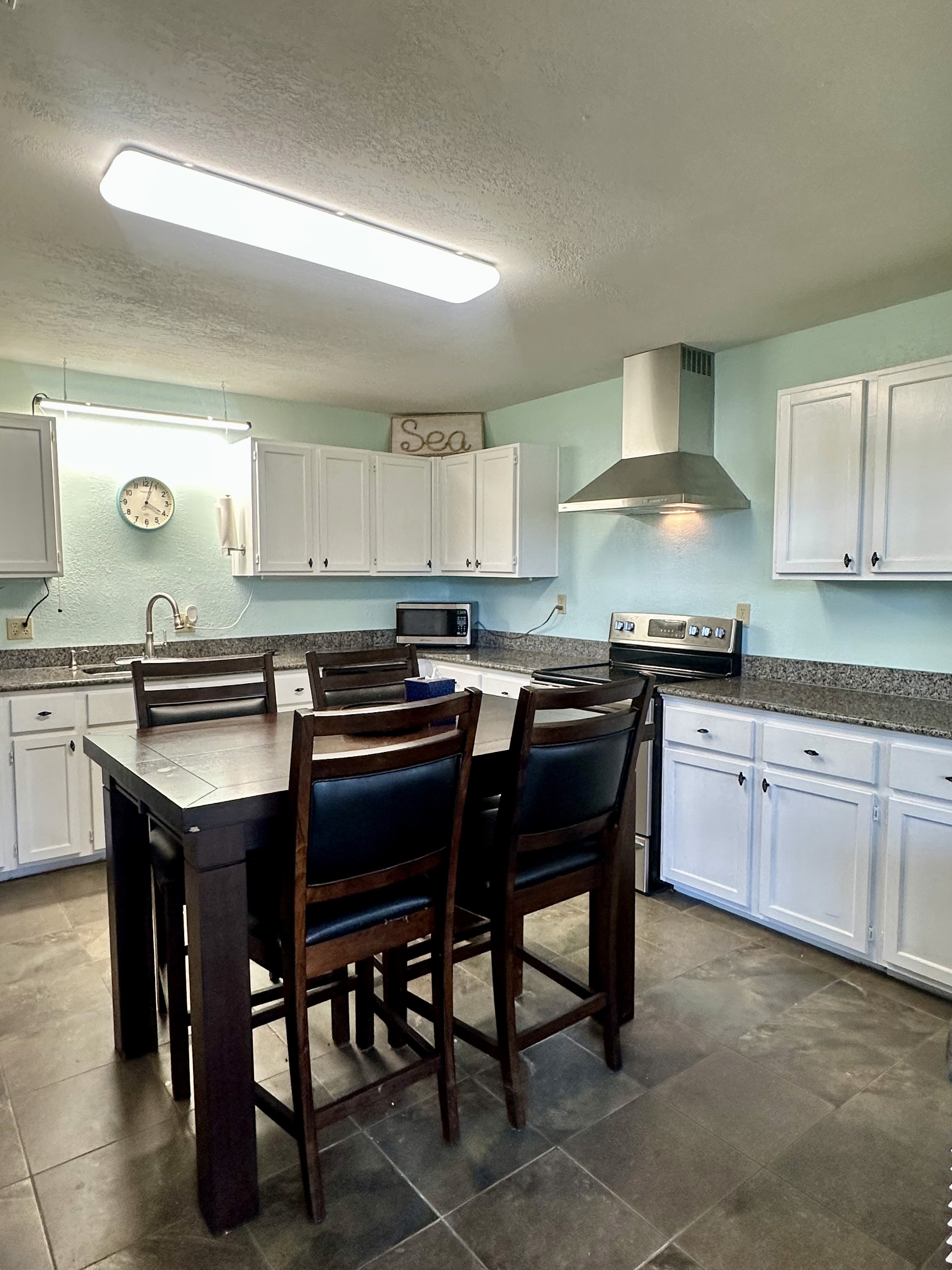 A spacious kitchen with white cabinets, granite countertops, a stainless steel range hood, and a large wooden dining table surrounded by dark chairs, illuminated by overhead lighting.