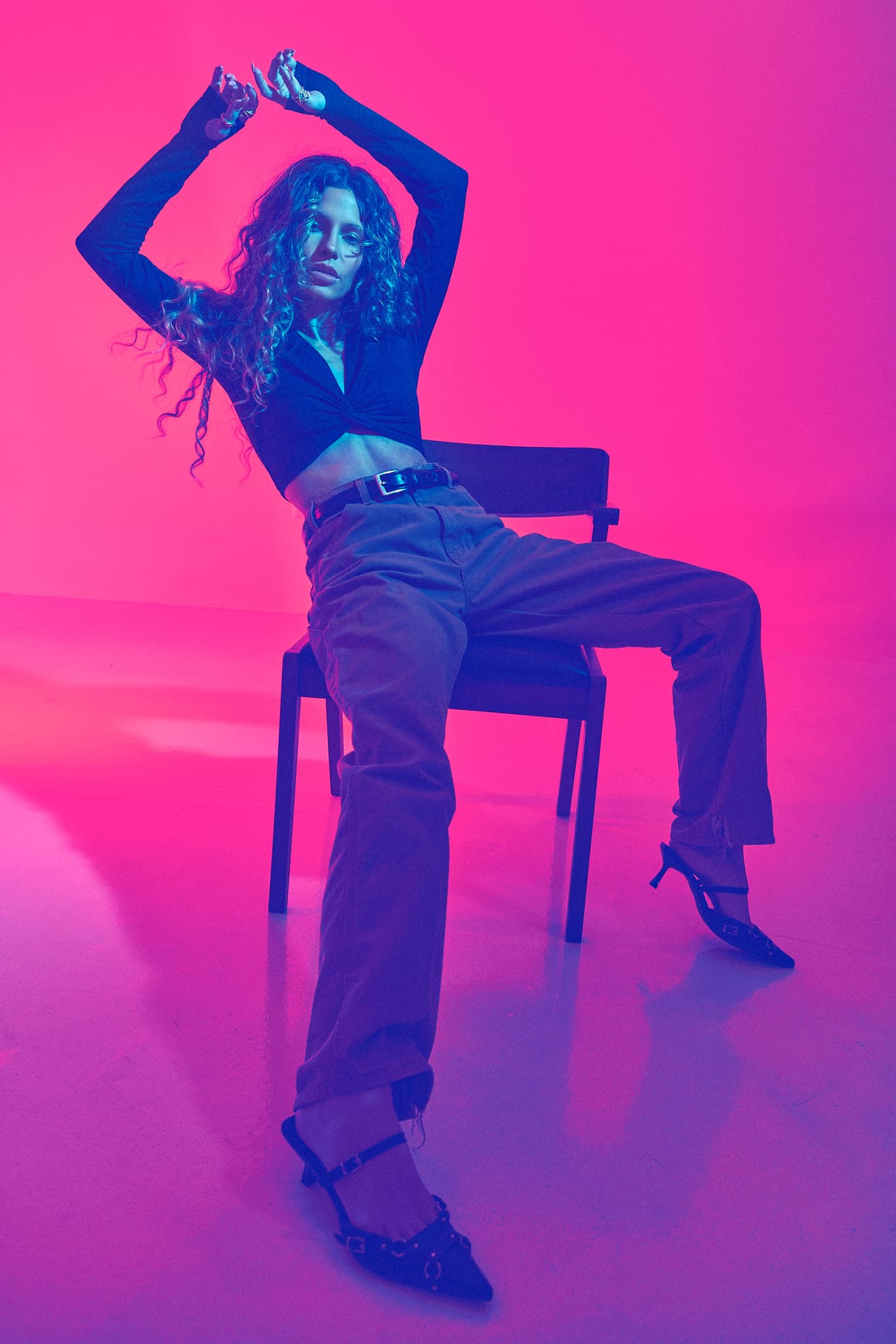 A model strikes a bold pose sitting in a chair with pink and purple lighting in a photography studio.