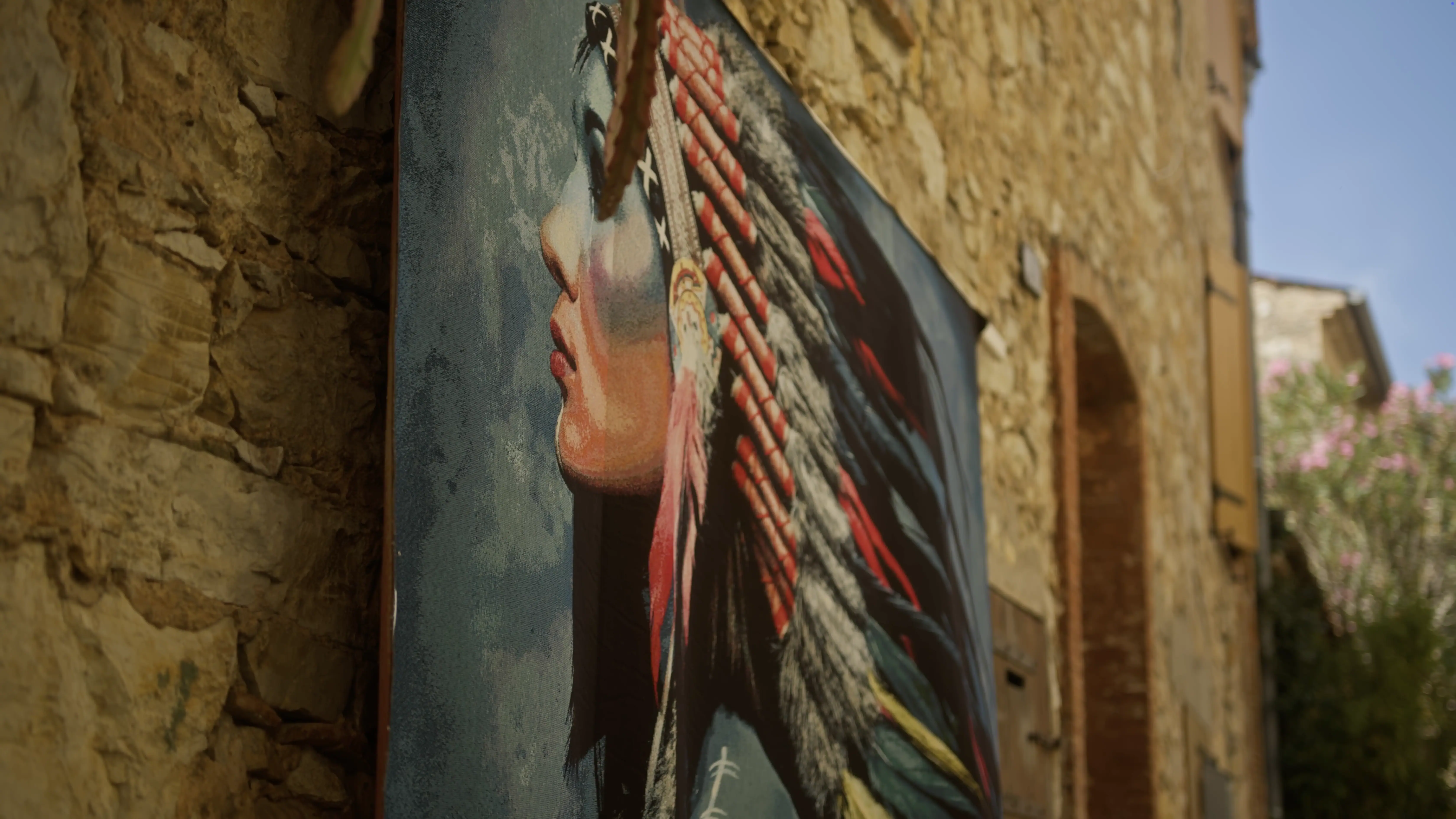 A large, colorful painting of a person wearing a traditional headdress is displayed against a rustic stone wall, with bright feathers and vibrant colors contrasting the texture of the wall.