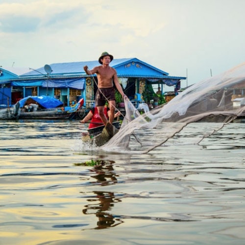 A man standing on a small boat throws a fishing net into the water with houses and boats in the background.
