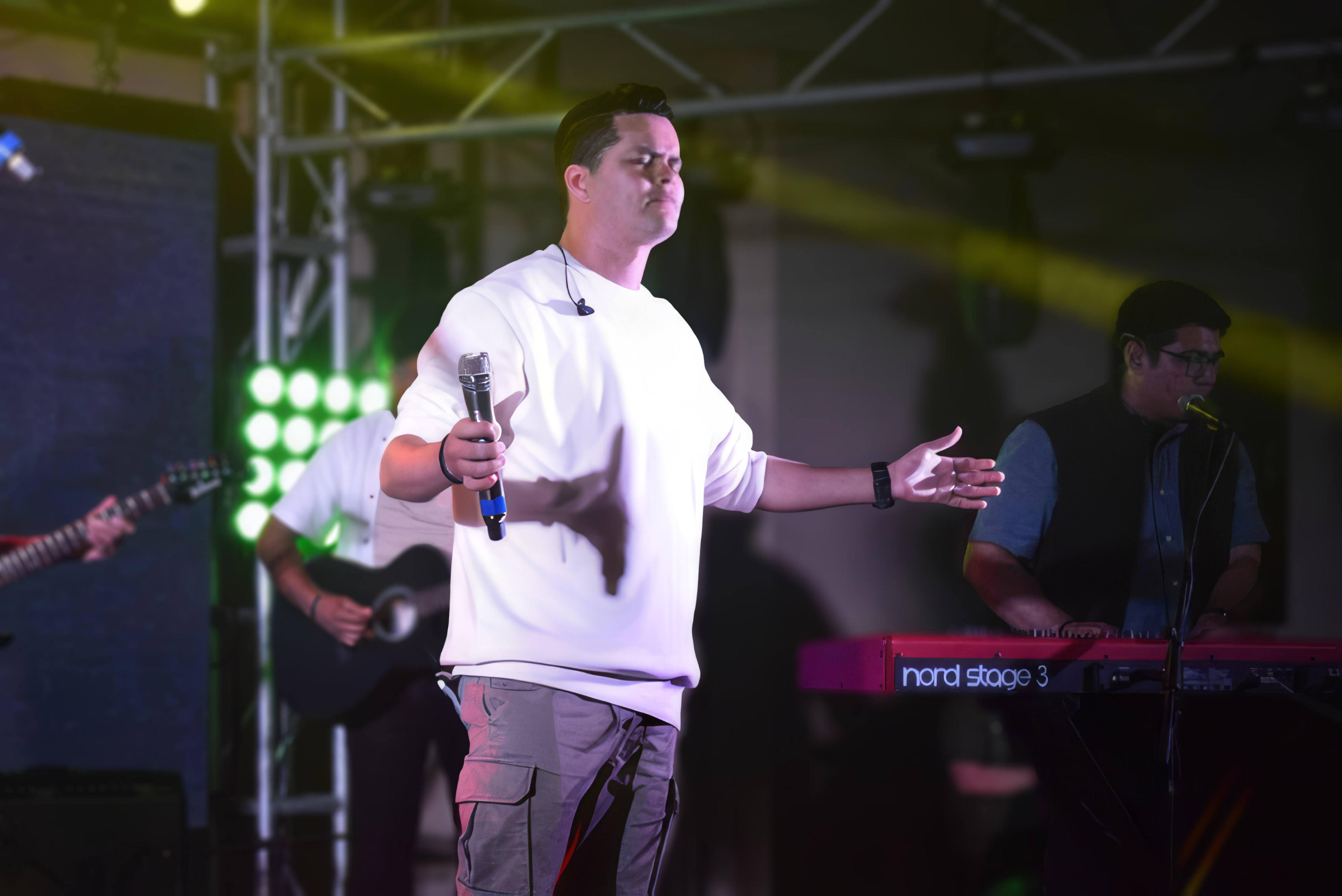 Dani Sierra performing in a white shirt gestures while singing on stage, with musicians playing in the background.