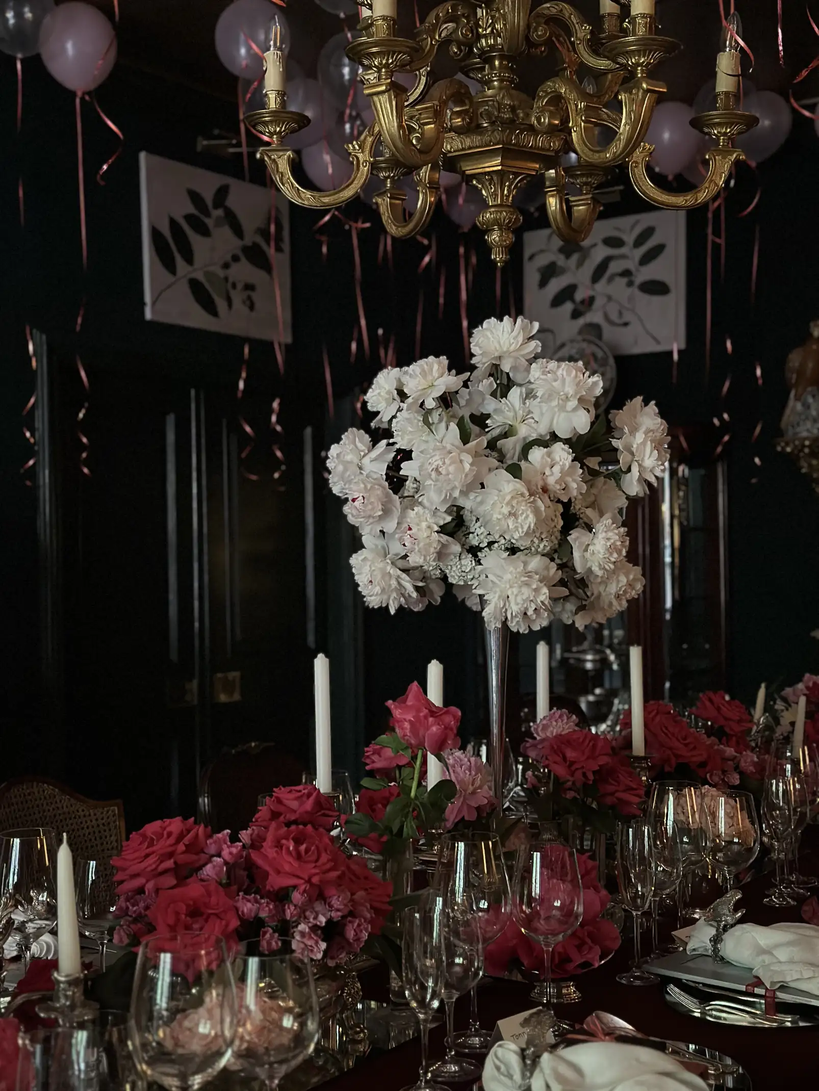 Mesa elegante decorada con centros florales en tonos blanco y fucsia, velas altas y copas de cristal bajo una lámpara de araña dorada, en un salón oscuro adornado con globos y cintas rosadas que cuelgan del techo.