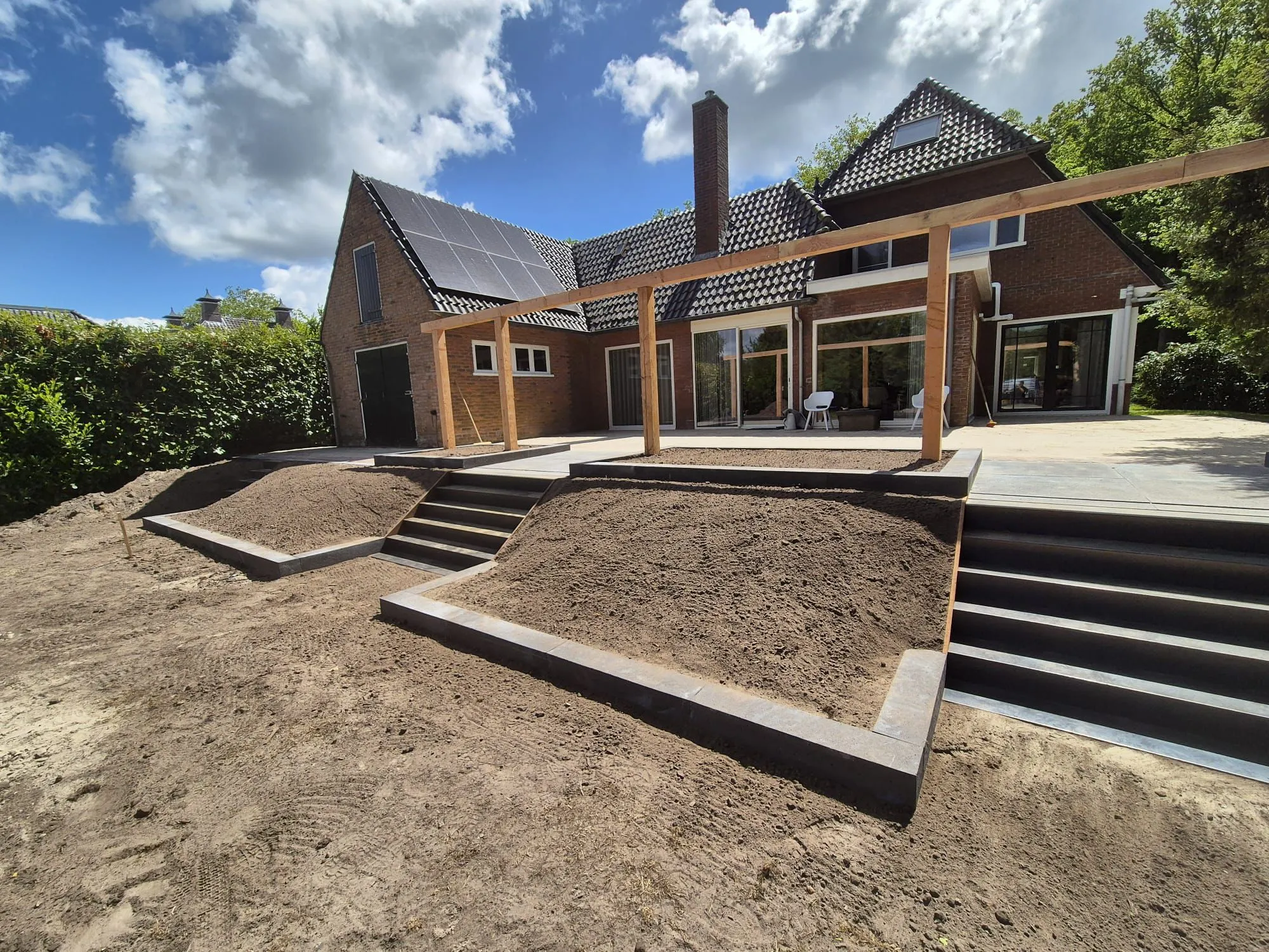 Rustiek terras met oud-Hollandse tegels, trap naar lager gelegen tuin en houten pergola bij woning in Oldeberkoop, klaar voor beplanting.
