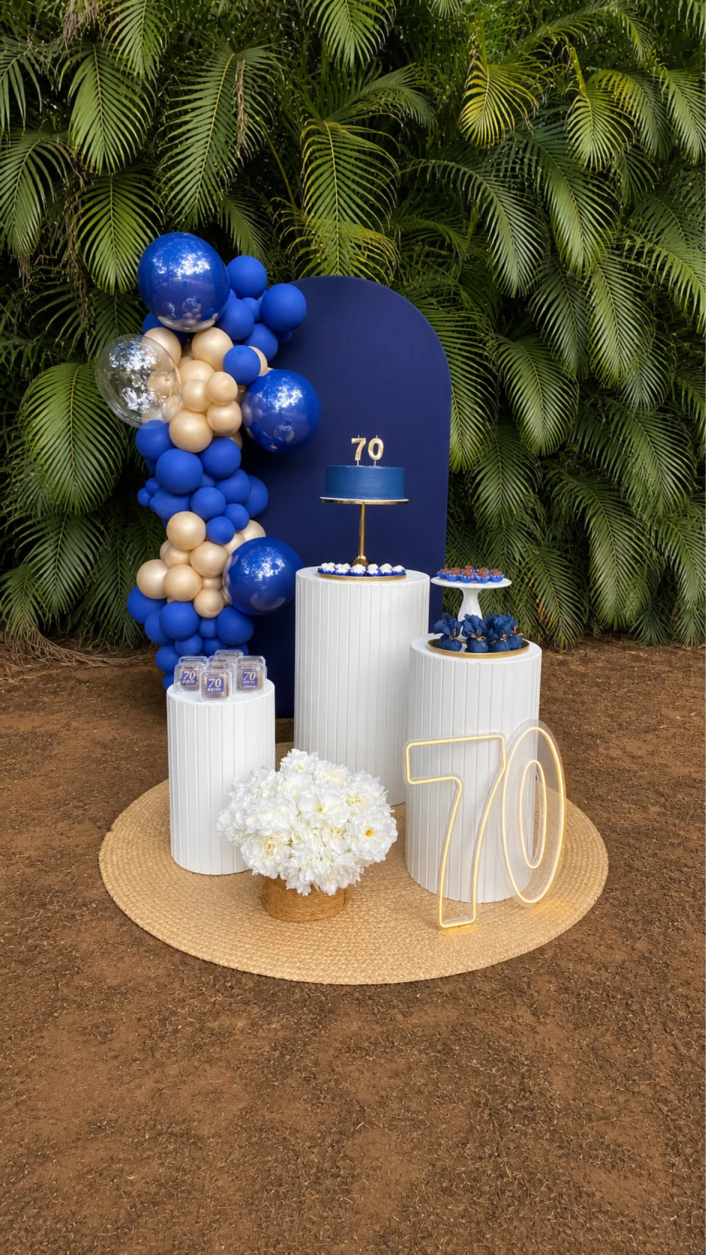 Decoração de aniversário em azul e dourado, com painel azul, arco de balões, cilindros brancos, flores e número iluminado.