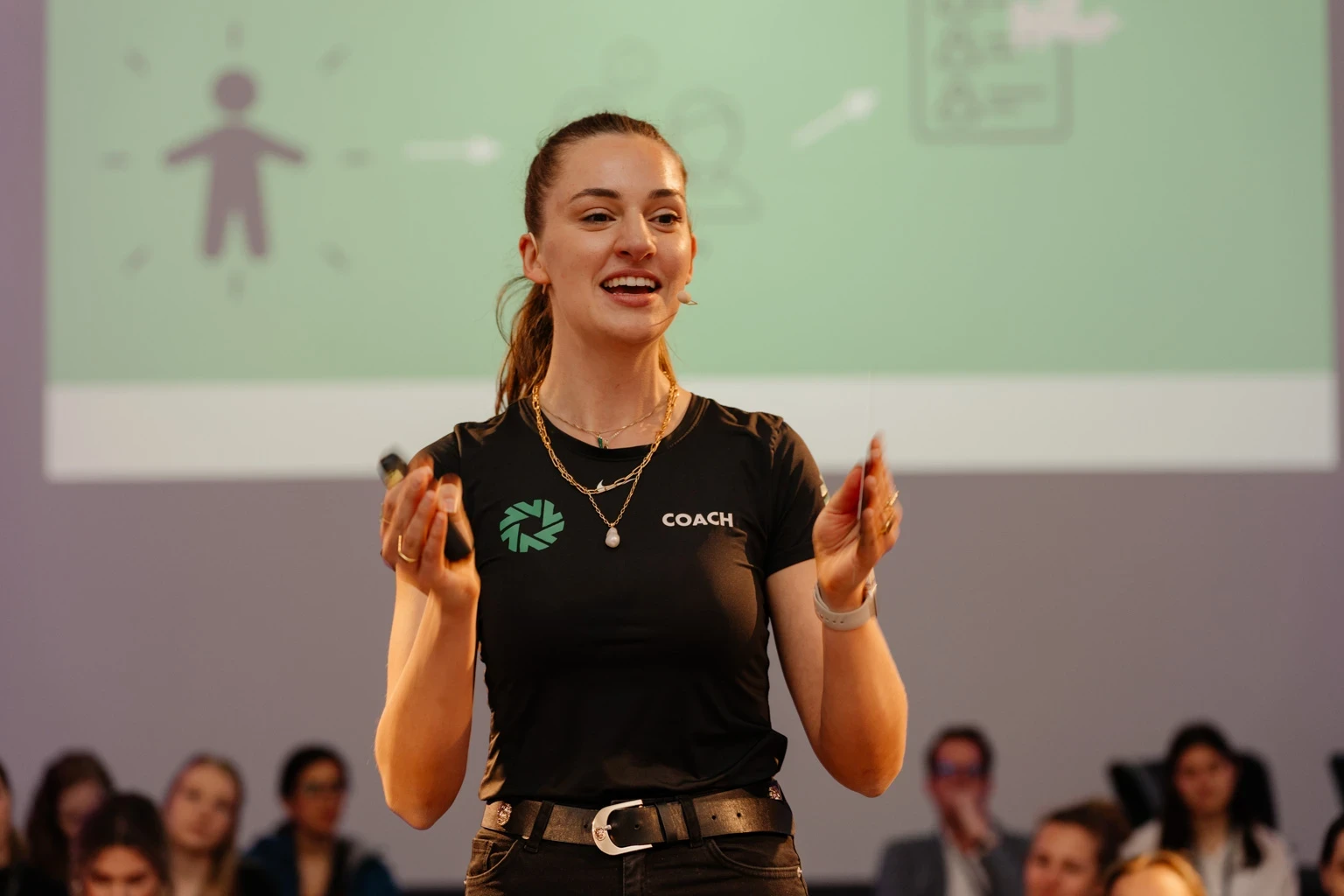 Eine junge Frau im Mittelpunkt, die gestikuliert. Im Hintergrund sind Menschen zu sehen, die gepannt zu ihr schauen. 