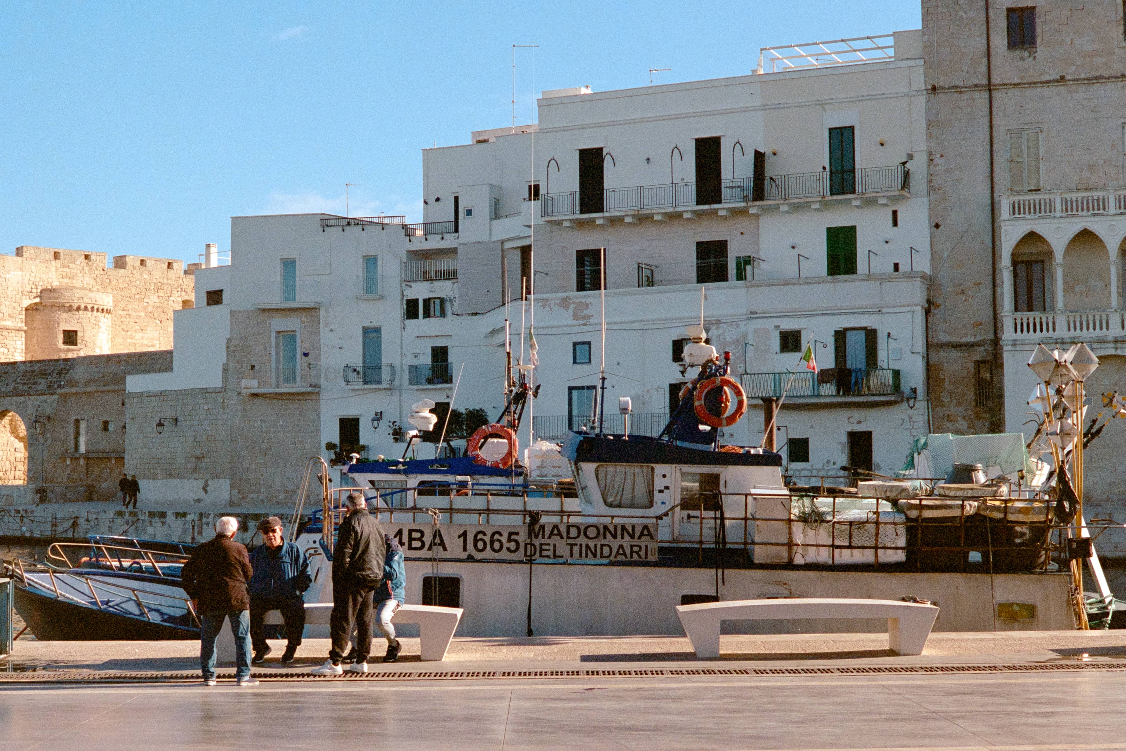 A group of people joyfully gather outdoors, featuring a woman in a white tank top smiling. The scene is lively, with colorful outfits and casual interactions.