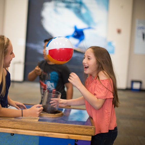 Dos chicas están jugando con un balón de playa flotante en una exhibición de ciencia, con una imagen borrosa de un surfista en el fondo.