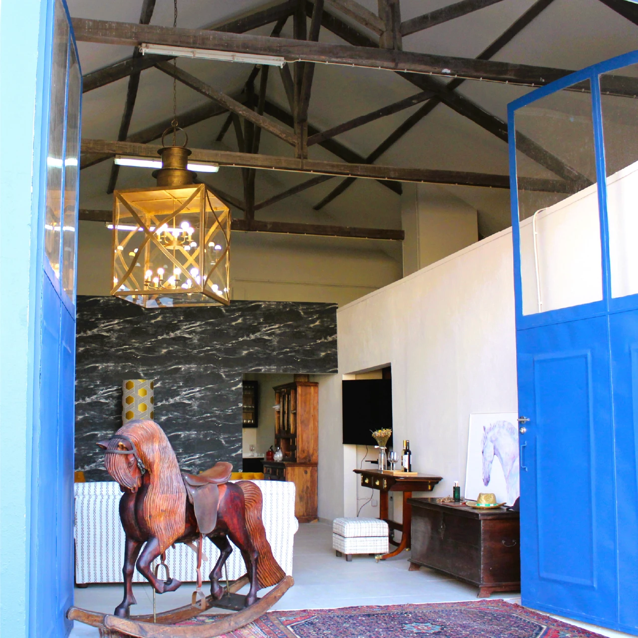 Open blue doors reveal a rustic interior featuring a wooden rocking horse, ornate chandelier, and a marble-patterned wall, creating a cozy atmosphere.