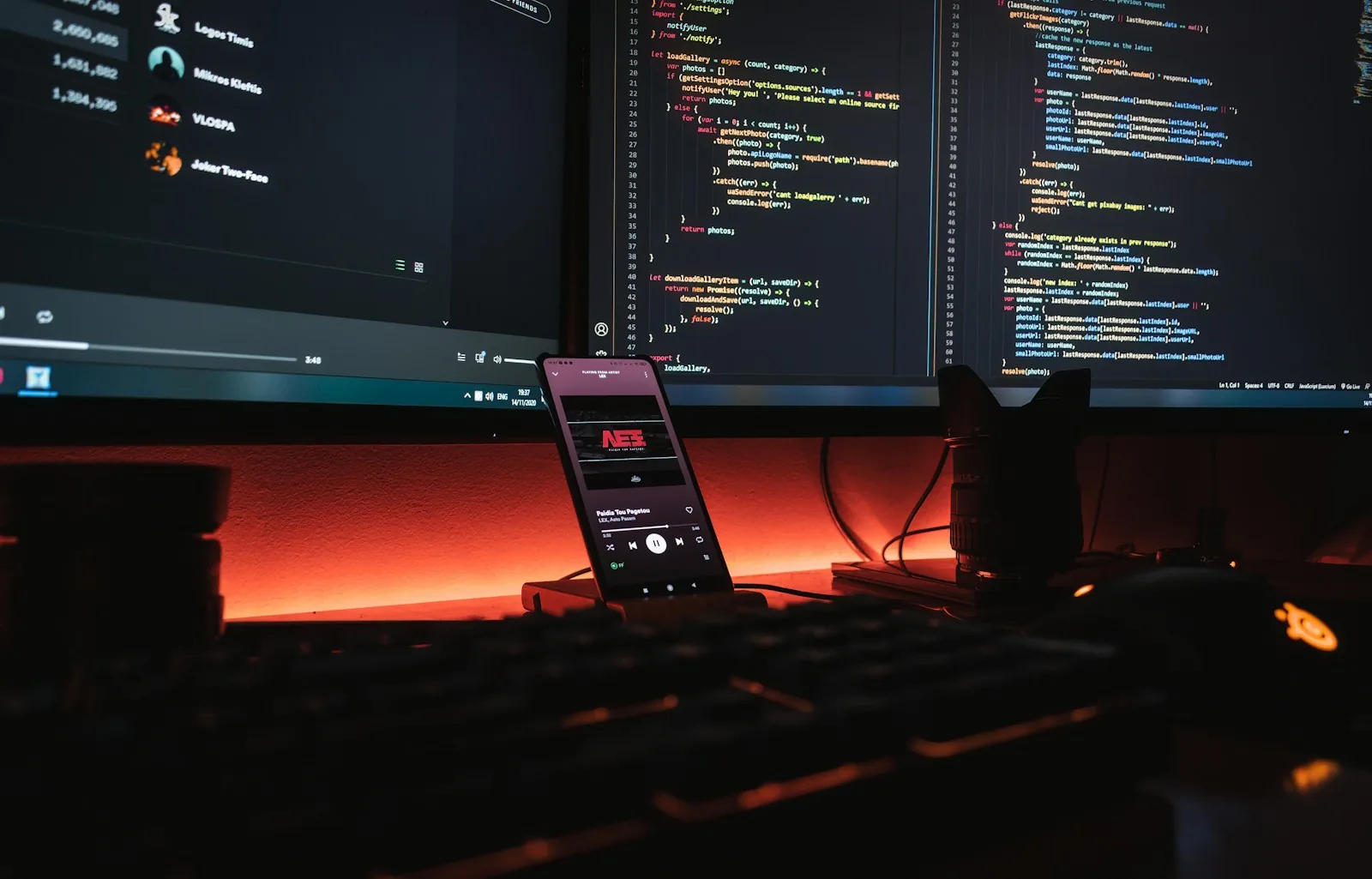 Smartphone running a music player app propped in front of two monitors showing JavaScript code, lit by an orange desk light