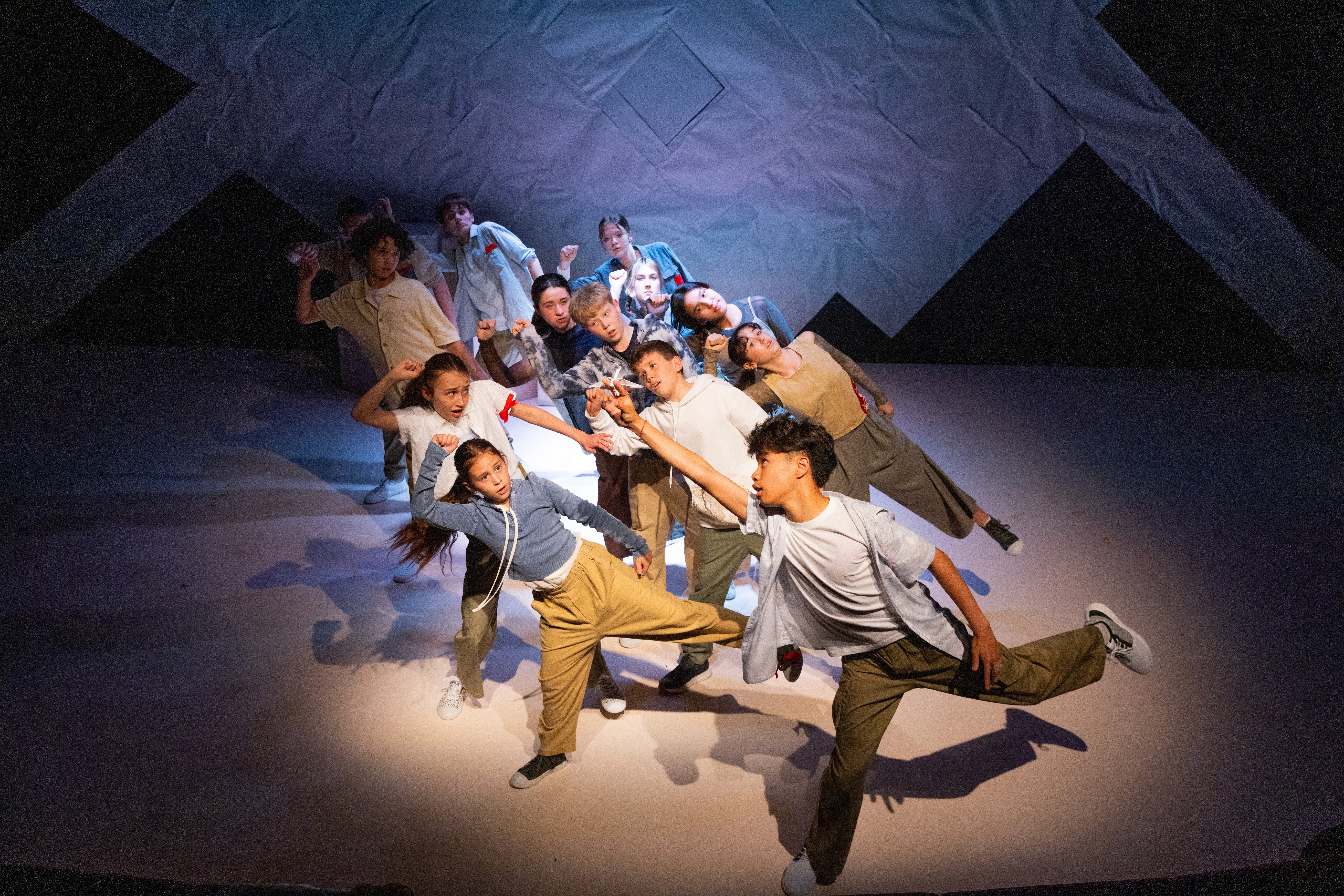 A large ensemble of young performers strike a stylised action pose mid-movement, arms extended and bodies angled in formation on a stark geometric set with soft, directional stage lighting.