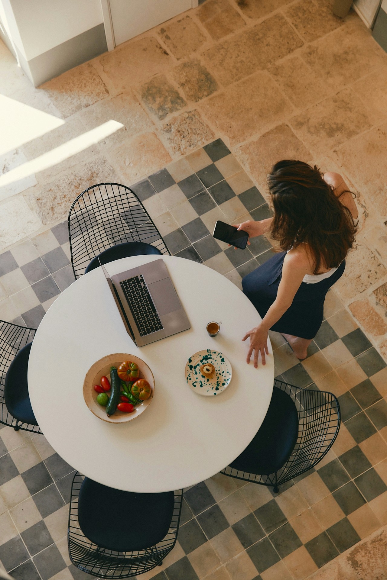 Vue plongeante d'une femme tenant un téléphone, près d'une table avec ordinateur, café, pâtisserie et légumes sur un sol carrelé.