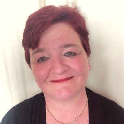 Uma pessoa com cabelo ruivo curto sorri calorosamente para a câmera, vestindo uma blusa escura, joias discretas e maquiagem leve, em um ambiente interno neutro.